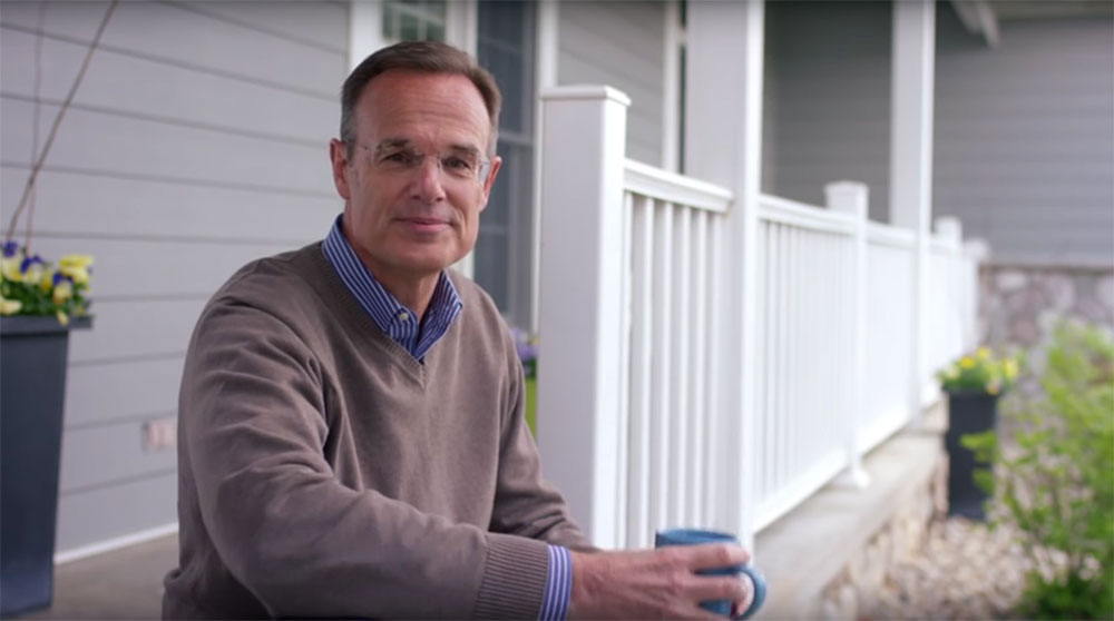 An older gentleman wearing a sweater and glasses sits on his front porch steps holding a blue mug and looks pleasantly at the camera. The porch is bright and full of spring flowers.