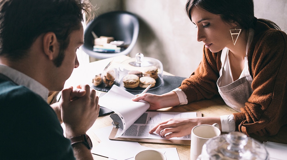 A young woman wearing an apron flips through paperwork on a clipboard across a wooden counter top from man. The counter has lots of baked goods and a cup of coffee. They are in her shop.