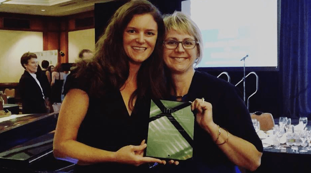 Two smiling women pose for the camera while holding a plaque together in a busy conference room event.
