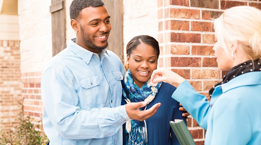 A young couple stand in each other's arms outside of a brick home while being handed a key from a businesswoman.