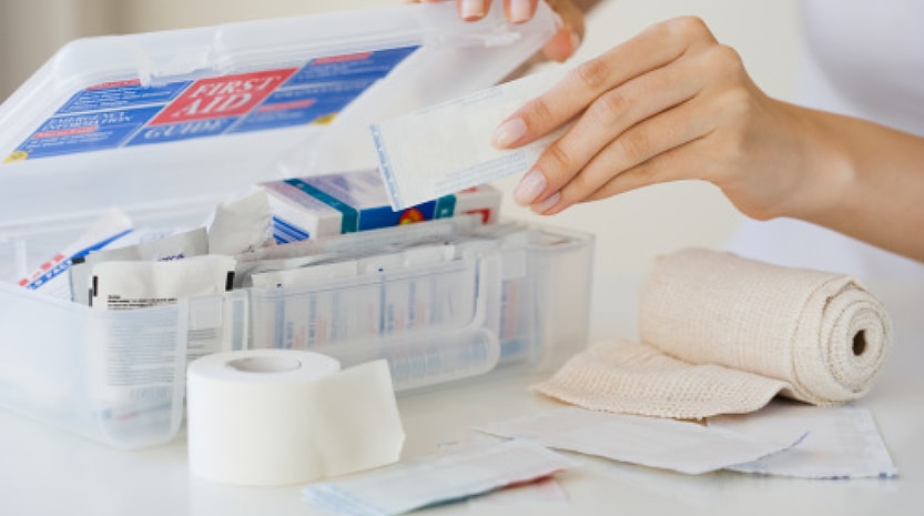 A woman holds open a first aid kit and is reaching to place bandages inside. Gauze and more items still sit on the table.