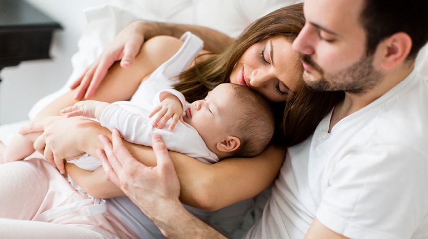 A woman holding a baby lays in her husbands arms in their pajamas on a bed.