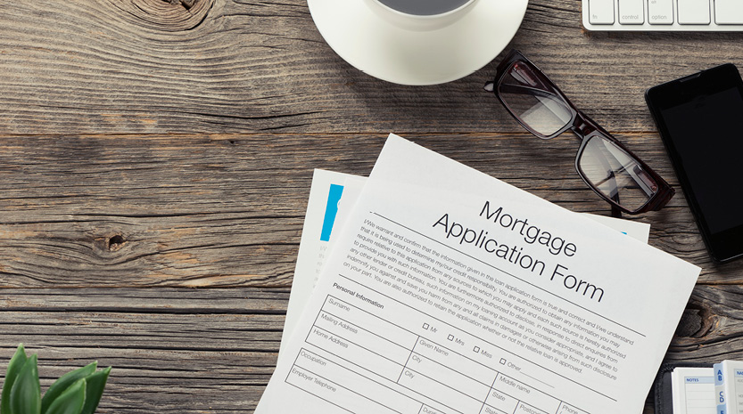 A photo of a cluttered wooden desktop containing a coffee cup, glasses, cell phone, computer keyboard, and a mortgage application form.