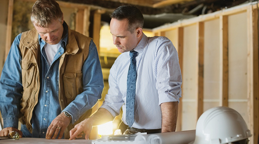 A construction worker and business lean over blueprints on a table within a building under construction.