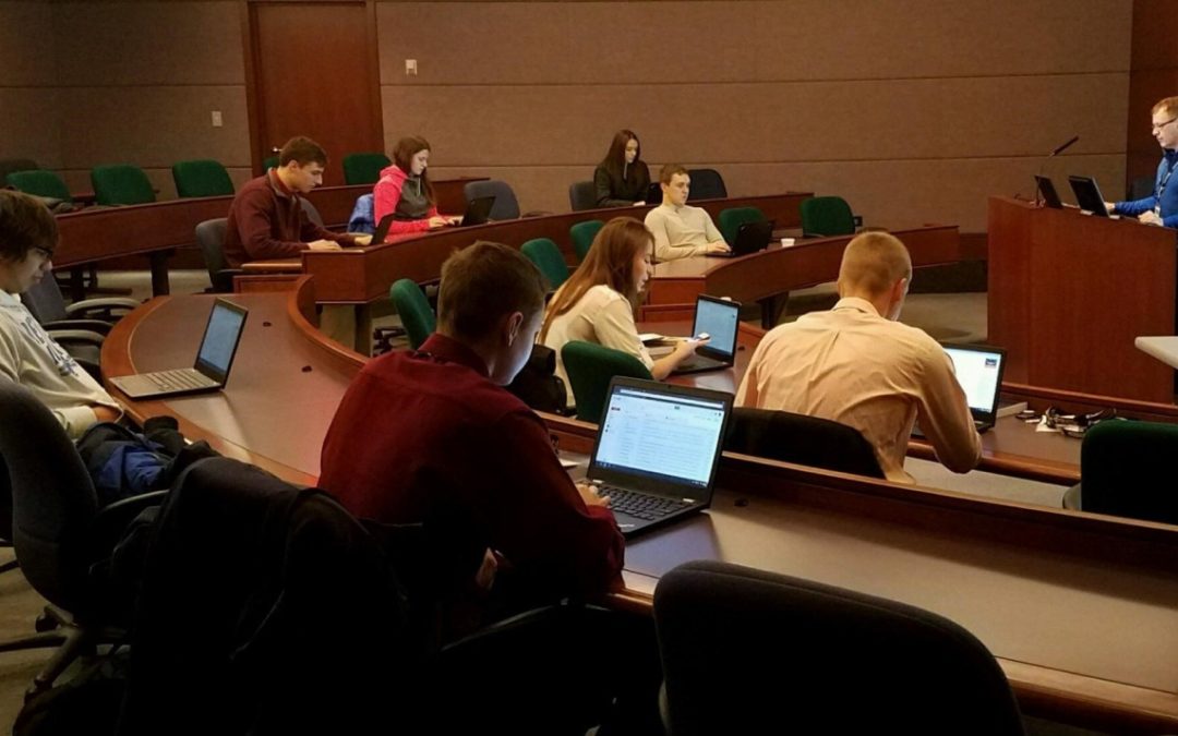 A group of young students work on laptops in a lecture hall while someone teaches at the front of the room.