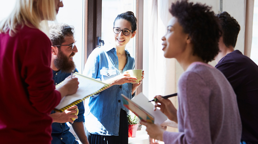 College students holding notebooks and coffee stand facing each other in a discussion group in front of a sunny window.