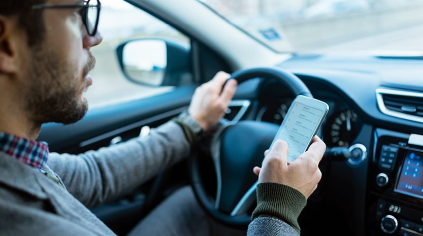 A young man with glasses sits in the driver seat of a car with one hand on the steering wheel and the other hand holding his phone.