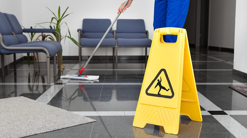 A janitor mops a tile floor lobby next to a yellow wet floor sign.