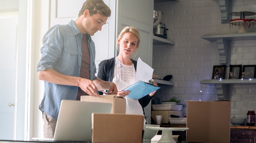 A man and a woman stand in a kitchen while putting labels on boxes and going through paperwork on a clipboard.