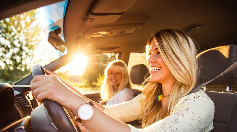 A mother and daughter smile while driving a car down a wooded road at sunset.