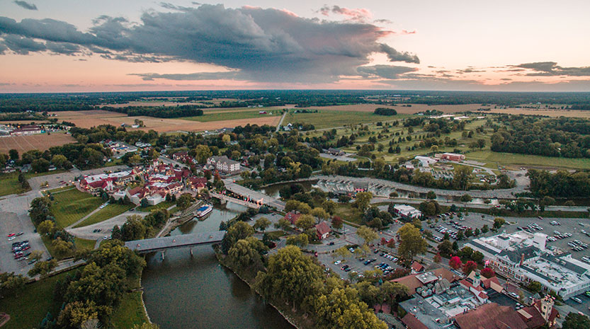 A beautiful aerial photo of downtown Frankenmuth at sunset. There are roads and shops and rooftops with a cloudy orange sunset in the distance.