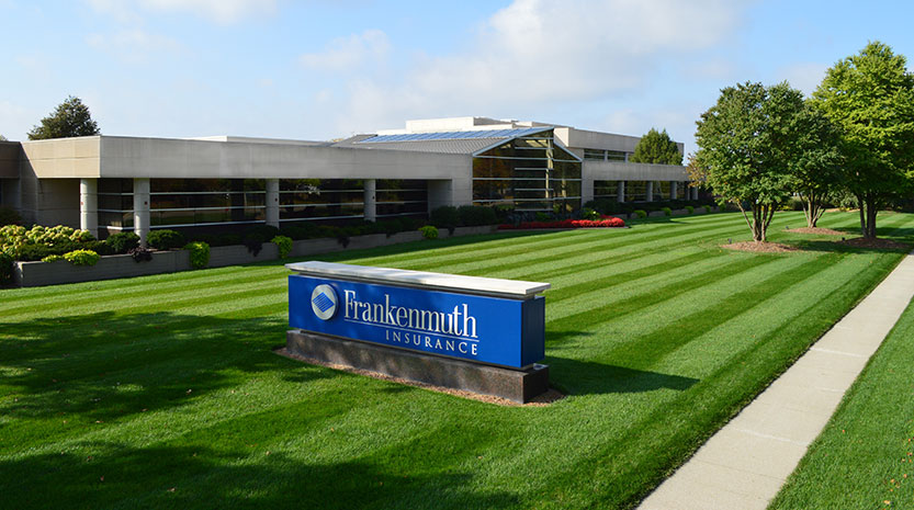 A cement and glass commercial building stands mightily behind a freshly mowed lawn on a sunny day. A blue Frankenmuth Insurance sign sits adjacent to the building next to a sidewalk.