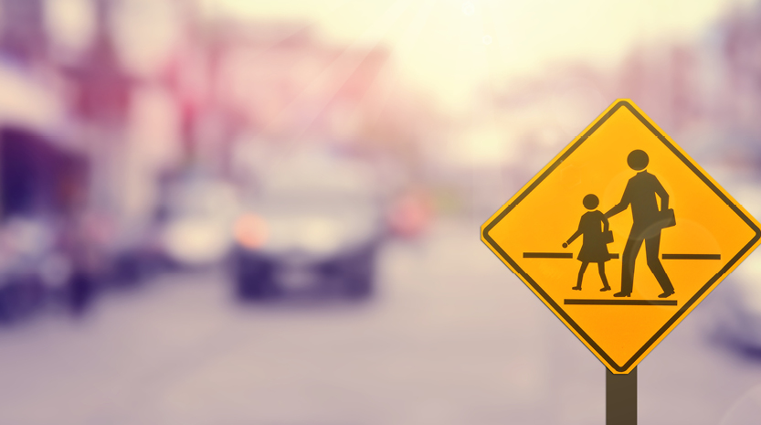 A yellow pedestrian crossing sign with a busy street in the background.