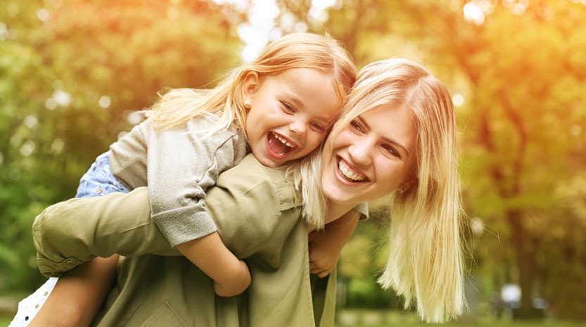 A mother leans over with her daughter on her back. They are both laughing on a sunny day outside.