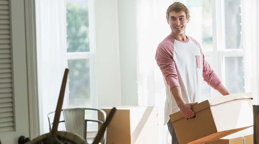 A man stands holding a large cardboard box in a sunny kitchen surrounded by other cardboard boxes on the floor.