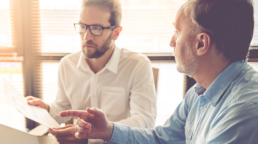 Two men sit at a table discussing life insurance paperwork in a bright room.