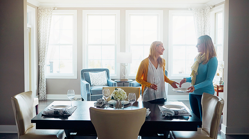 Two women talk and smile as they are setting the kitchen table.